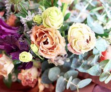 A bouquet bursting with seasonal flowers and foliage in shades of coral, tan, lilac and purple with lots of seasonal greenery and foliages and loads of texture.
