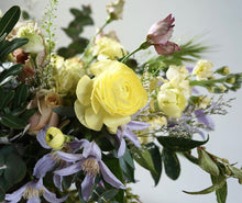 A hand-tied bouquet bursting with seasonal flowers and foliage in shades of yellow and lilac with lots of seasonal greenery and foliages and loads of texture