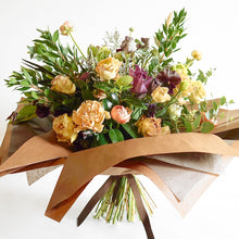 A bouquet bursting with seasonal flowers and foliage in shades of coral, tan, lilac and purple with lots of seasonal greenery and foliages and loads of texture.